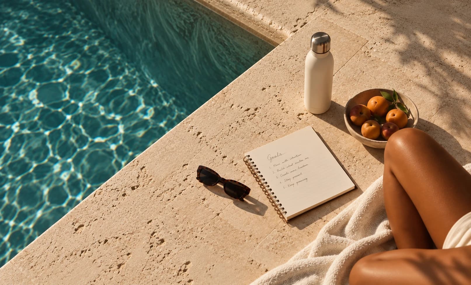 A notebook, fruit and water bottle beside a sunlit pool